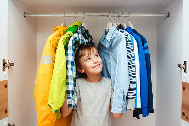 A light skinned male presenting child is seen center frame. He’s standing in a closet surrounded by from the left a yellow raincoat a green windbreaker and a white black green and blue plaid button front shirt. To his left, frame right, or a light blue oxford cloth button front shirt a blue white and gray striped sweater a Carolina blue shirt with a white stripe a royal blue fleece. He is wearing a short sleeved gray T-shirt. He has dark hair that sweeps across his forehead. He is looking up toward right frame apparently at a blue shirt that makes him very happy.