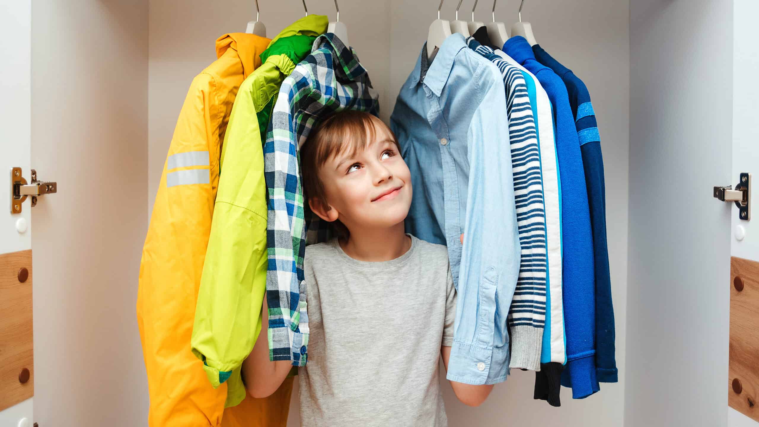 A light skinned male presenting child is seen center frame. He’s standing in a closet surrounded by from the left a yellow raincoat a green windbreaker and a white black green and blue plaid button front shirt. To his left, frame right, or a light blue oxford cloth button front shirt a blue white and gray striped sweater a Carolina blue shirt with a white stripe a royal blue fleece. He is wearing a short sleeved gray T-shirt. He has dark hair that sweeps across his forehead. He is looking up toward right frame apparently at a blue shirt that makes him very happy.