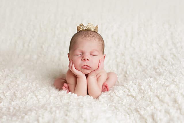 A newborn baby sits on a white blanket held up by its hands and elbows while wearing a gold crown