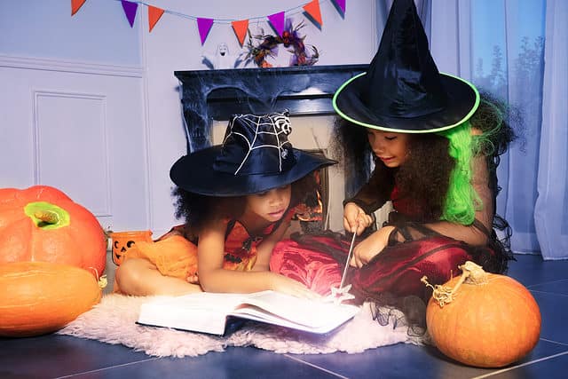 Two girls read wizard book in Halloween witch costumes cast a spell with magic wand holding sit by fireplace