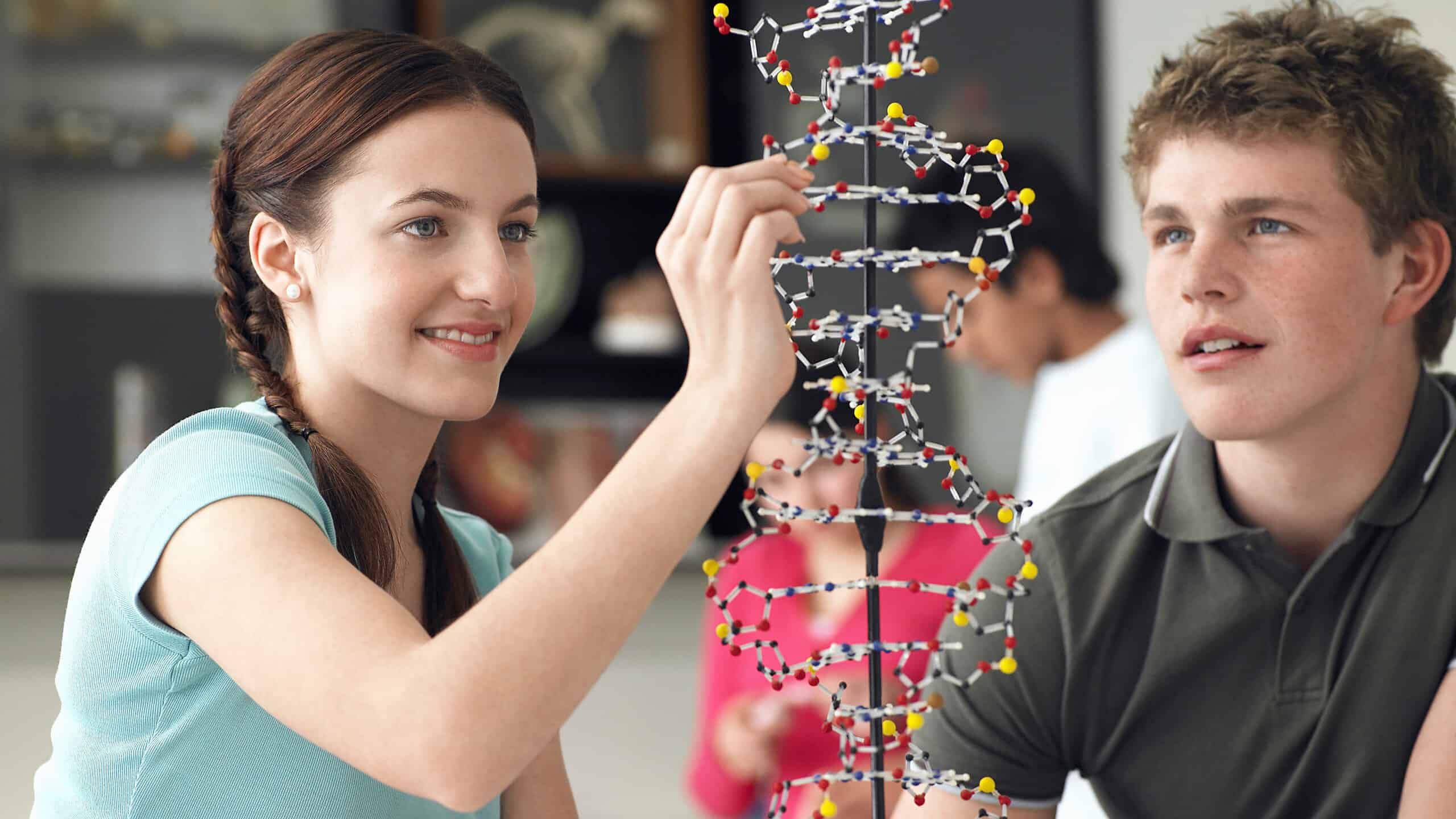 Happy teenage students examining DNA models and taking notes in science class