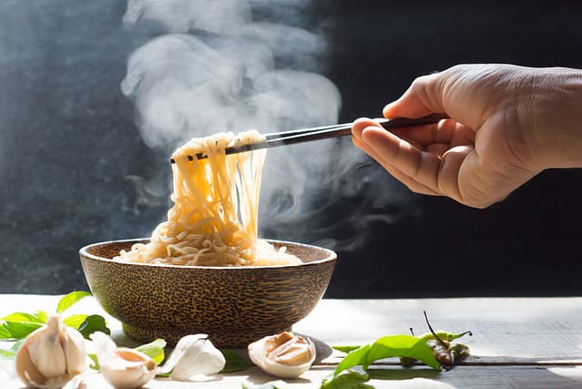 Hand uses chopsticks to pickup tasty noodles with steam and smoke in bowl on wooden background, selective focus. Asian meal on a table, junk food concept