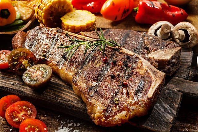 Close up of a succulent tender grilled porterhouse steak seasoned with pepper and rosemary on a wooden board with fresh halved tomatoes, mushrooms, corncobs and bell peppers