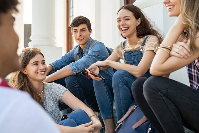 Group of happy young friends sitting in college campus and talking. Cheerful group of smiling girls and guys feeling relaxed after university exam. Excited millenials laughing and having fun outdoor.