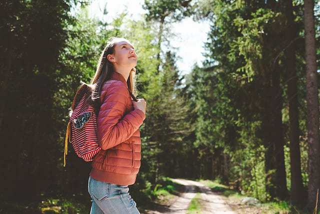 Teen tourist girl admiring forest, red puffer jacket and orange hiking backpack.