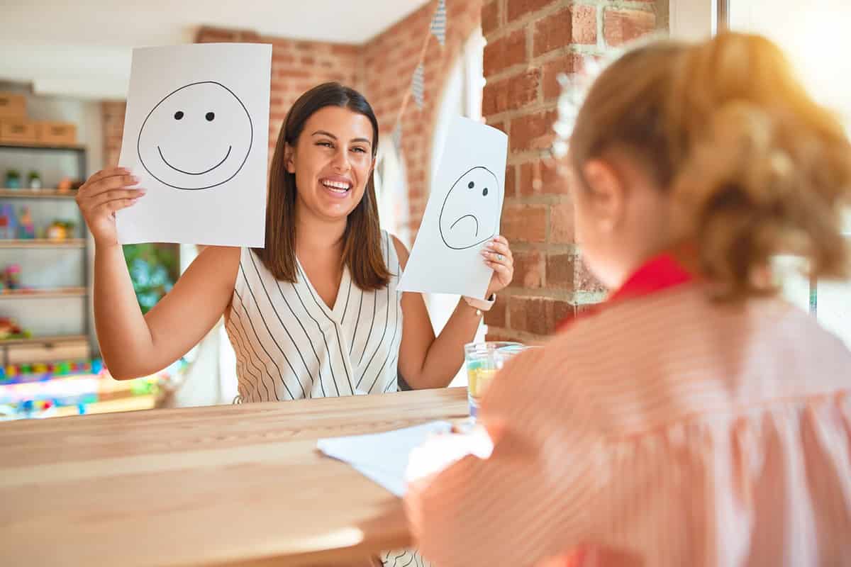 Beautiful psycologist and blond toddler girl wearing school uniform sitting on desk doing therapy using emoji emotions at consulting room