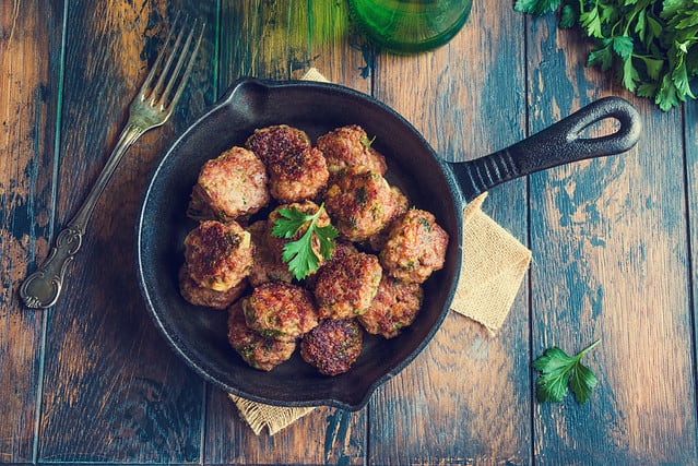 Homemade roasted beef meatballs in cast-iron skillet on wooden table in kitchen, fresh parsley, vintage fork, top view.