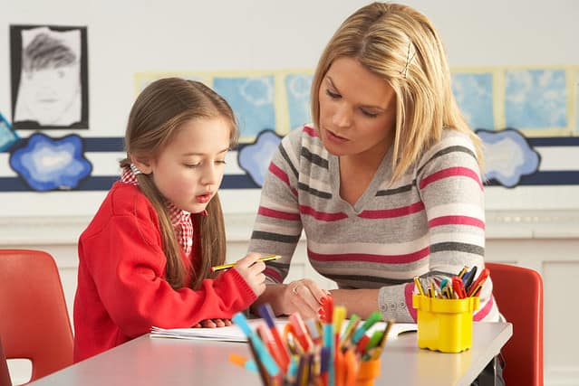 Female Primary School Pupil And Teacher Working At Desk In Classroom