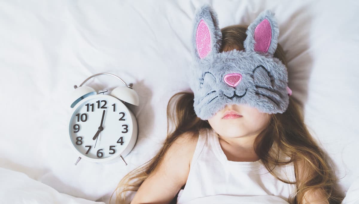 Little girl sleeping in bed under white blanket wearing grey bunny plush sleep mask with alarm clock on pillow. Early morning wake up. Putting kid to sleep.Mom's correct daily routine, rest for child.