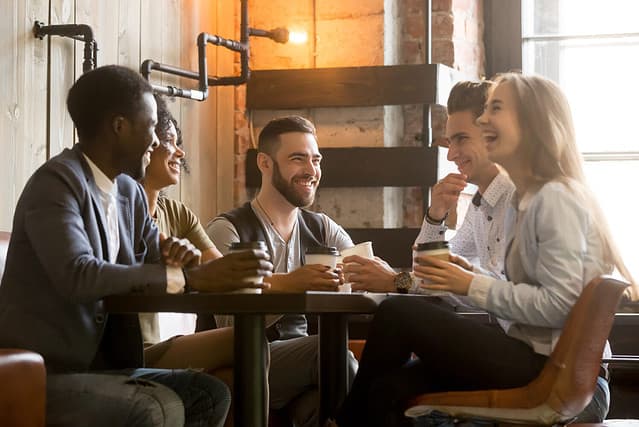 Multiracial friends having fun and laughing drinking coffee in coffeehouse, diverse young people talking joking sitting together at cafe table, multi ethnic millennials spending time in coffee shop