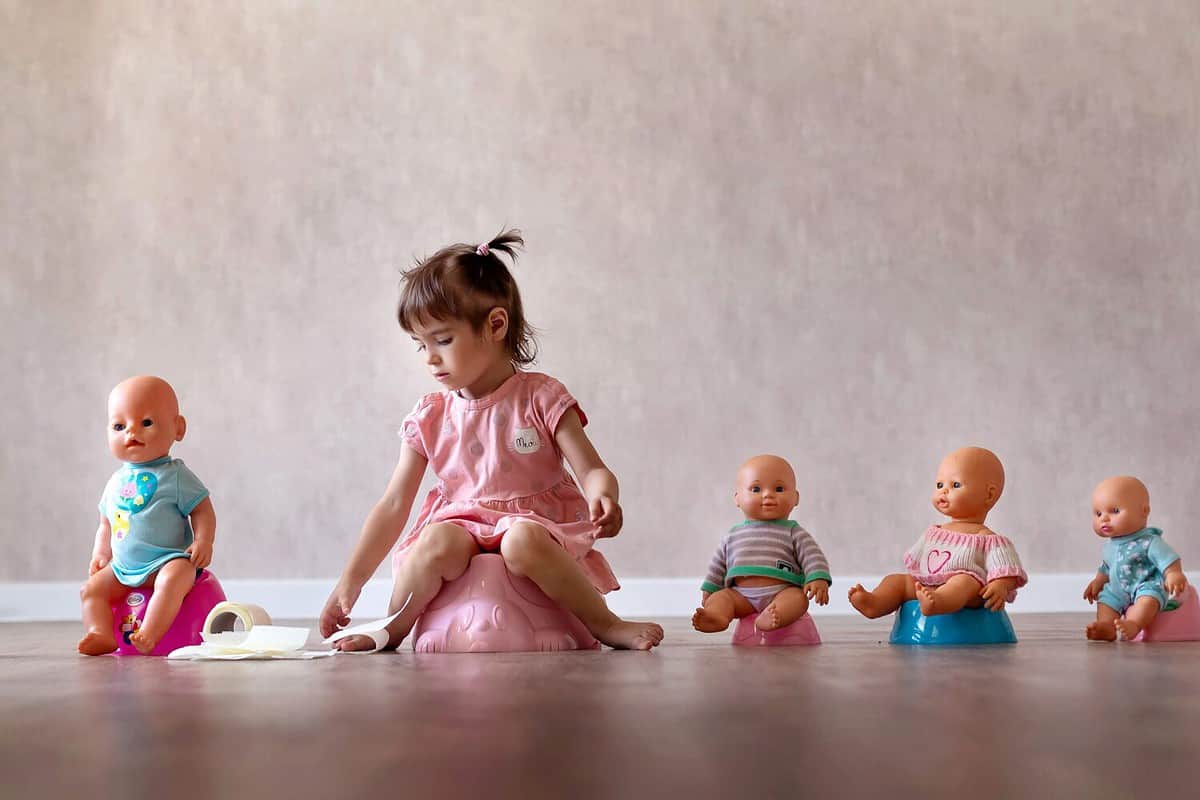 Beautiful smiling little baby sitting on potty. Cute adorable funny child girl using chamber pot together with her baby doll. Toilet training concept. Toddler learning to use the Toilet.