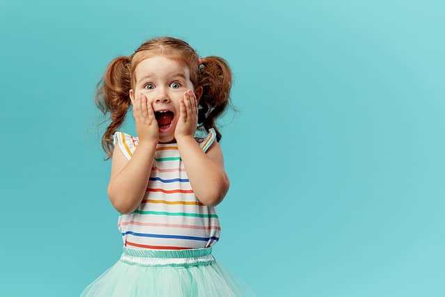 Portrait of surprised cute little toddler girl child standing isolated over blue background. Looking at camera. hands near open mouth