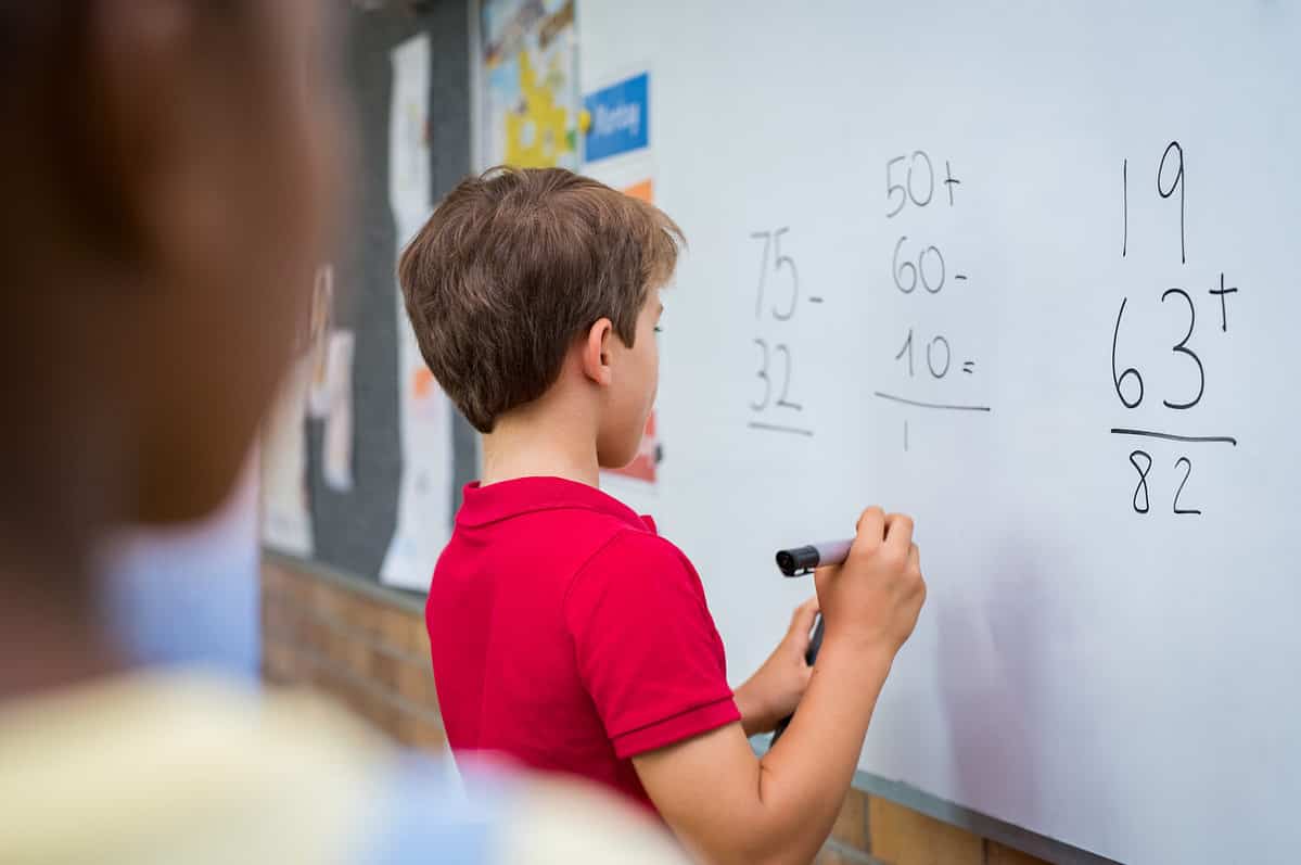 Rear view of young boy solving addition and subtraction on white board at school. Schoolboy thinking while solving math's sum. Child writing the solution of the mathematical operation in classroom.