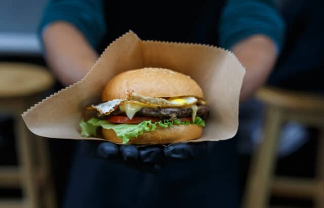 Fresh burger cooked at barbecue outdoors in craft paper. Cookout american bbq. Big hamburger with steak meat and vegetables closeup with chef unfocused at background. Street food, fast food.