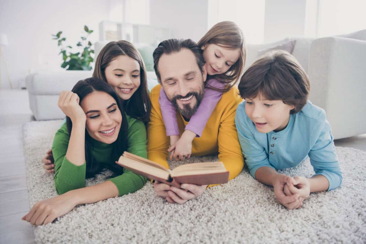 Cozy dreamy people enjoy free time lie carpet floor dad daddy read book mom mommy three small preteen kids listen in house indoors
