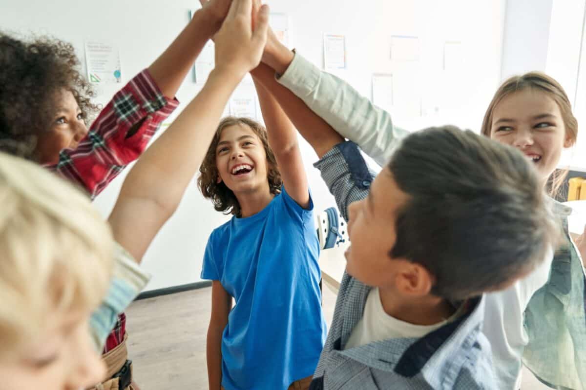 Happy diverse multiethnic kids junior school students group giving high five together in classroom. Excited children celebrating achievements, teamwork, diversity and friendship with highfive concept.