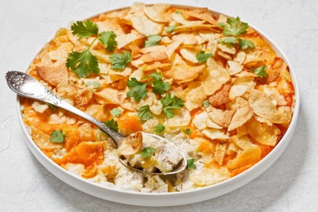 creamy cheesy chicken almond casserole topped with potato chips in baking dish with spoon on white concrete table, american recipe, horizontal view, close-up