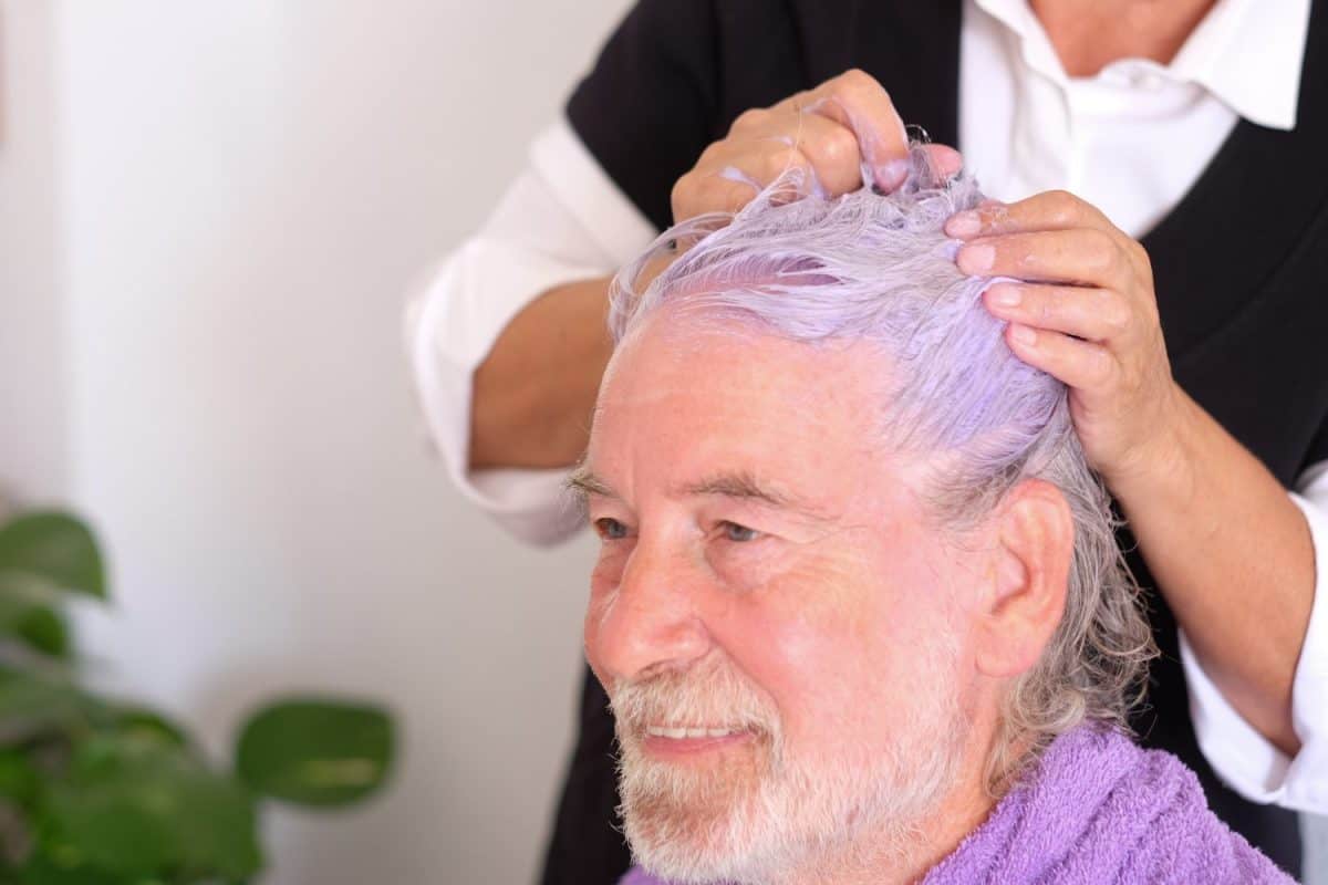 An elderly handsome man with white and grey hair and beard confident in the hairdresser. Looks at himself in the mirror. A violet special shampoo helps to remove yellow color from the hairs