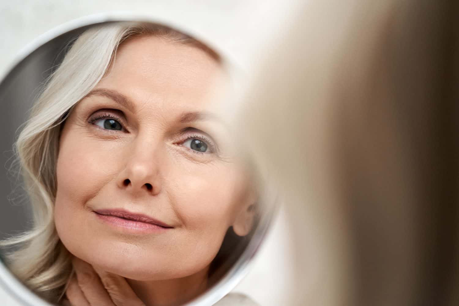 Happy 50s middle aged woman model touching face skin looking in mirror reflection. Smiling mature old lady pampering, healthy moisturized skin care, aging beauty, skincare treatment cosmetics concept.