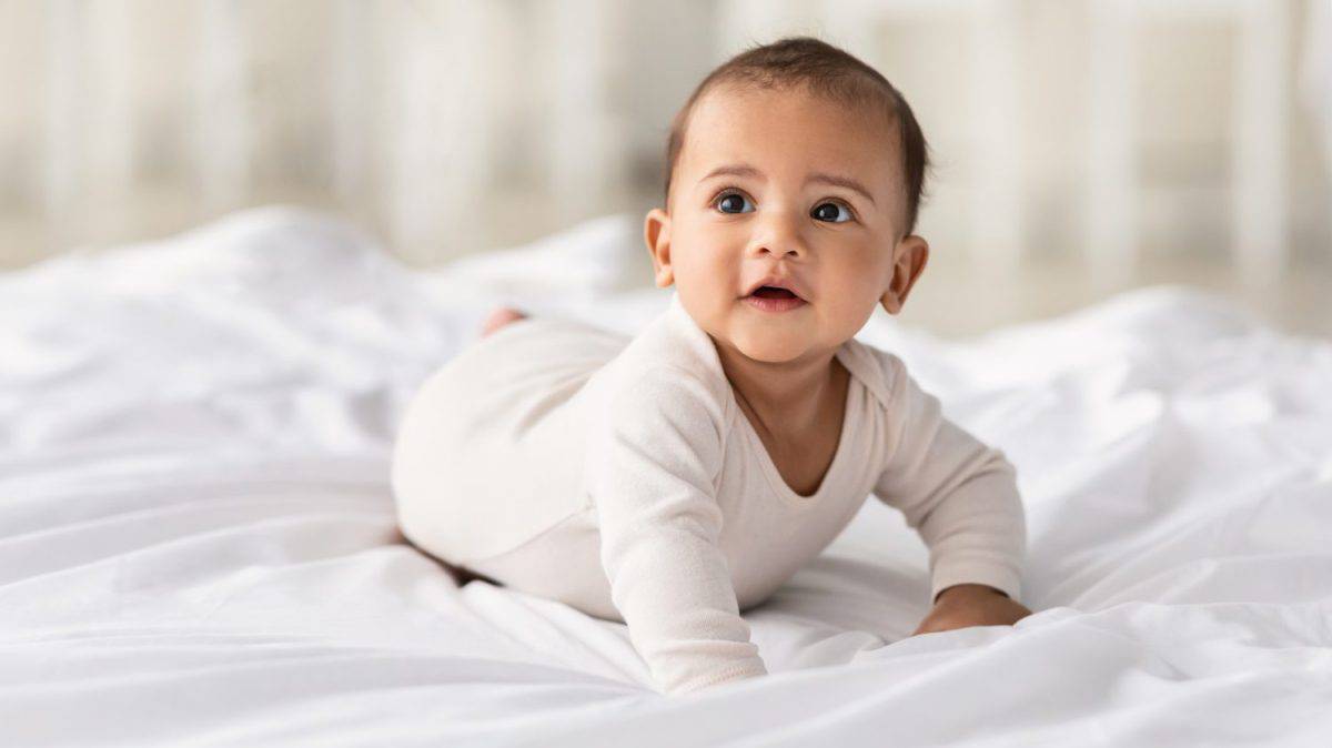 Childcare Concept. Portrait of cute little African American baby wearing bodysuit lying on white beedsheets at home. Black infant child crawling on bed in the bedroom. Selective focus, free copy space