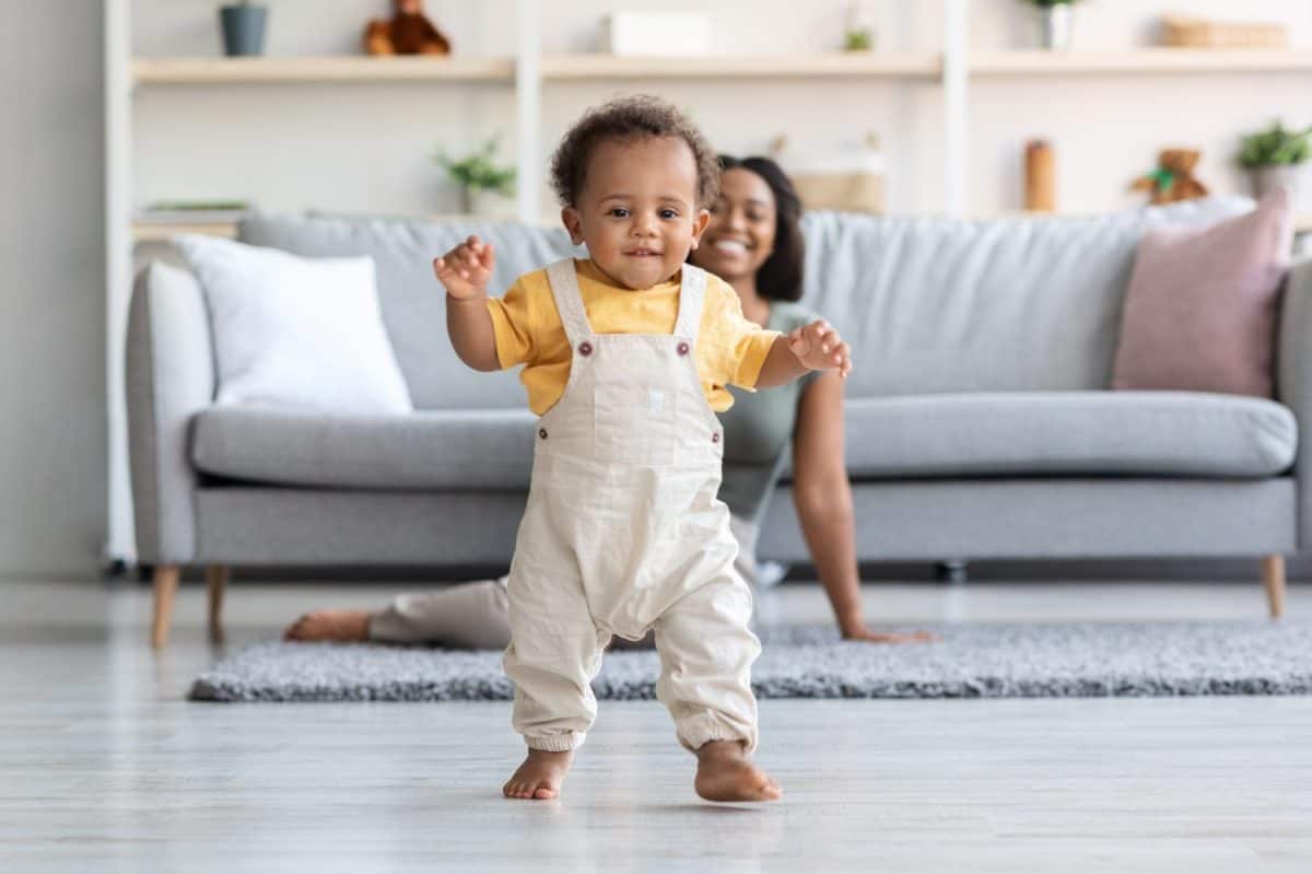 Baby Development. Cute Little Black Infant Boy Making First Steps At Home, Adorable African American Toddler Child Walking In Living Room, His Happy Mom Smiling On Background, Copy Space