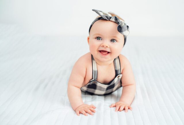 Seven month old baby child sitting on bed. Cute smiling little infant girl on white soft blanket. girl wearing headband. Charming blue eyed baby. Copy space.