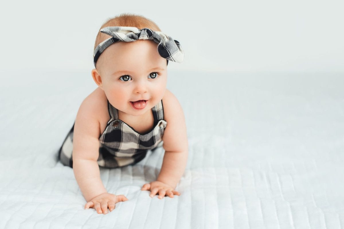 Seven month old baby child sitting on bed. Cute smiling little infant girl on white soft blanket. girl wearing headband. Charming blue eyed baby. Copy space.