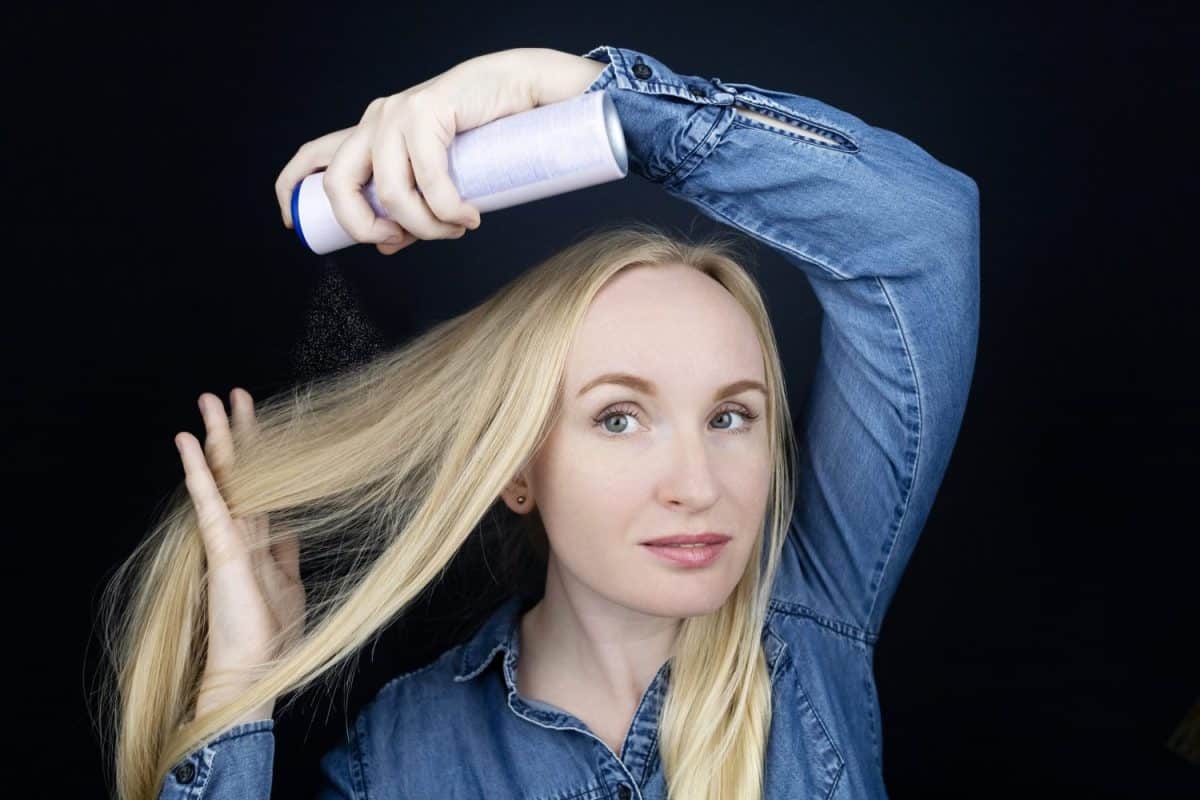 Dry shampoo. Blonde girl sprays shampoo on her hair. The problem of oily hair while traveling. An emergency remedy for excessive sebum production. Make your head clean without water. Shampoo for oily hair.