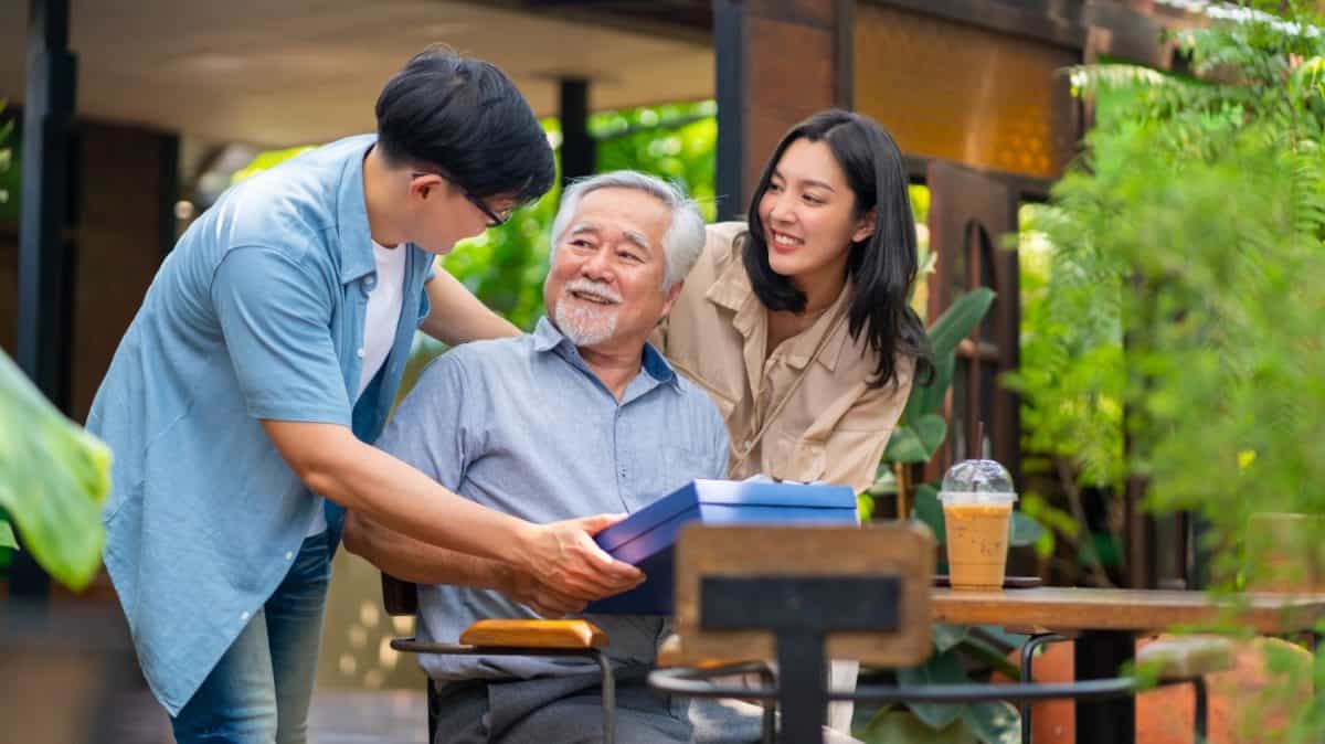 Asian couple surprised elderly father with Birthday gift at outdoor cafe restaurant on summer holiday vacation. Family relationship, celebrating father's day and older people health care concept.