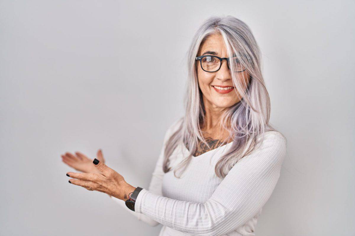 Middle age woman with grey hair standing over white background inviting to enter smiling natural with open hand. Shampoo for gray hair