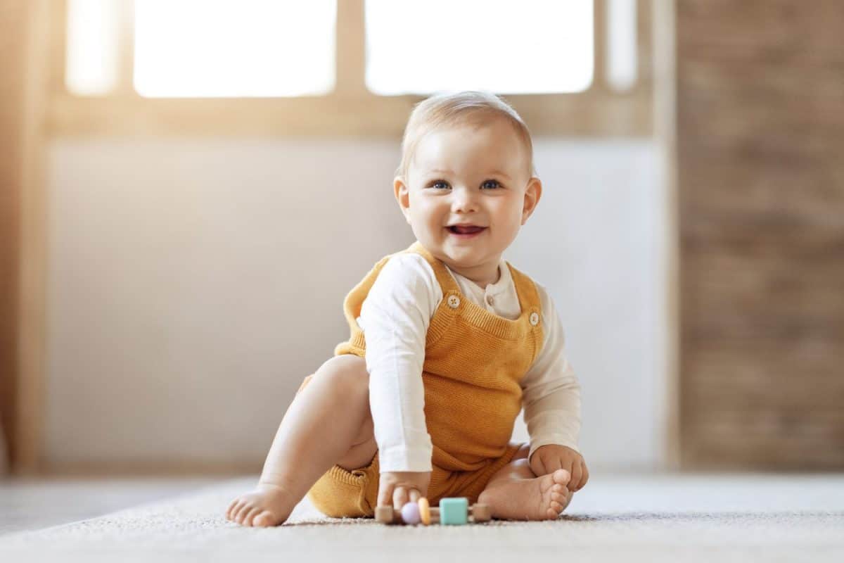 Adorable happy blonde infant baby playing with kids toys at home while sitting on carpet floor in living room. Portrait of smiling cute child toddler using colorful toys, copy space