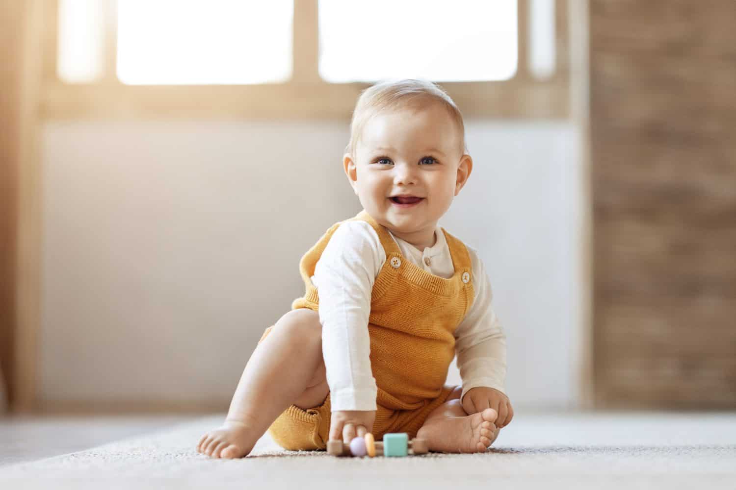 Adorable happy blonde infant baby playing with kids toys at home while sitting on carpet floor in living room. Portrait of smiling cute child toddler using colorful toys, copy space