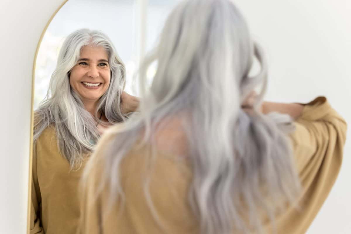 Happy positive elderly Hispanic woman mirror reflection and back view. Cheerful senior model touching long naturally grey hair, looking at herself, laughing, smiling