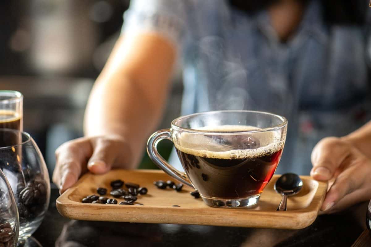 black drip coffee in glass cup, Barista making drip coffee by pouring spills hot water on coffee bean. Barista serve holding cup of hot black coffee or americano for serve on wooden table cafe shop