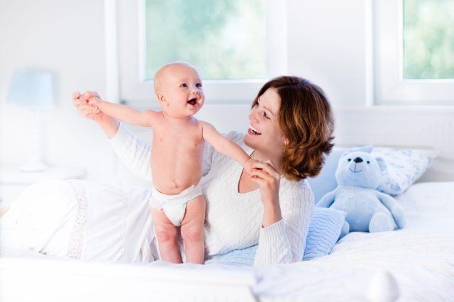 Mother and child on a white bed. Mom and baby boy in diaper playing in sunny bedroom. Parent and little kid relaxing at home. Family having fun together. Bedding and textile for infant nursery. These are the traits you got from your mother