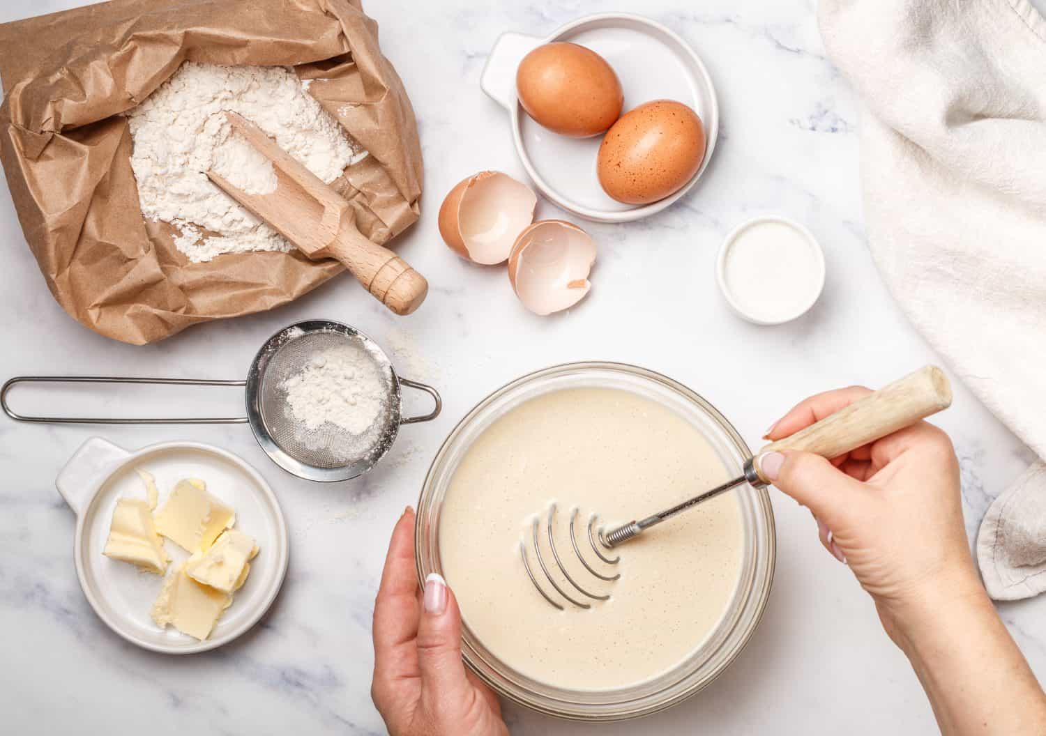 Woman prepares dough for homemade pancakes for Breakfast. Whisk for whipping in hands. Ingredients on the table - wheat flour, eggs, butter, sugar, salt, milk. Selective focus. How to make sheet pan pancakes.