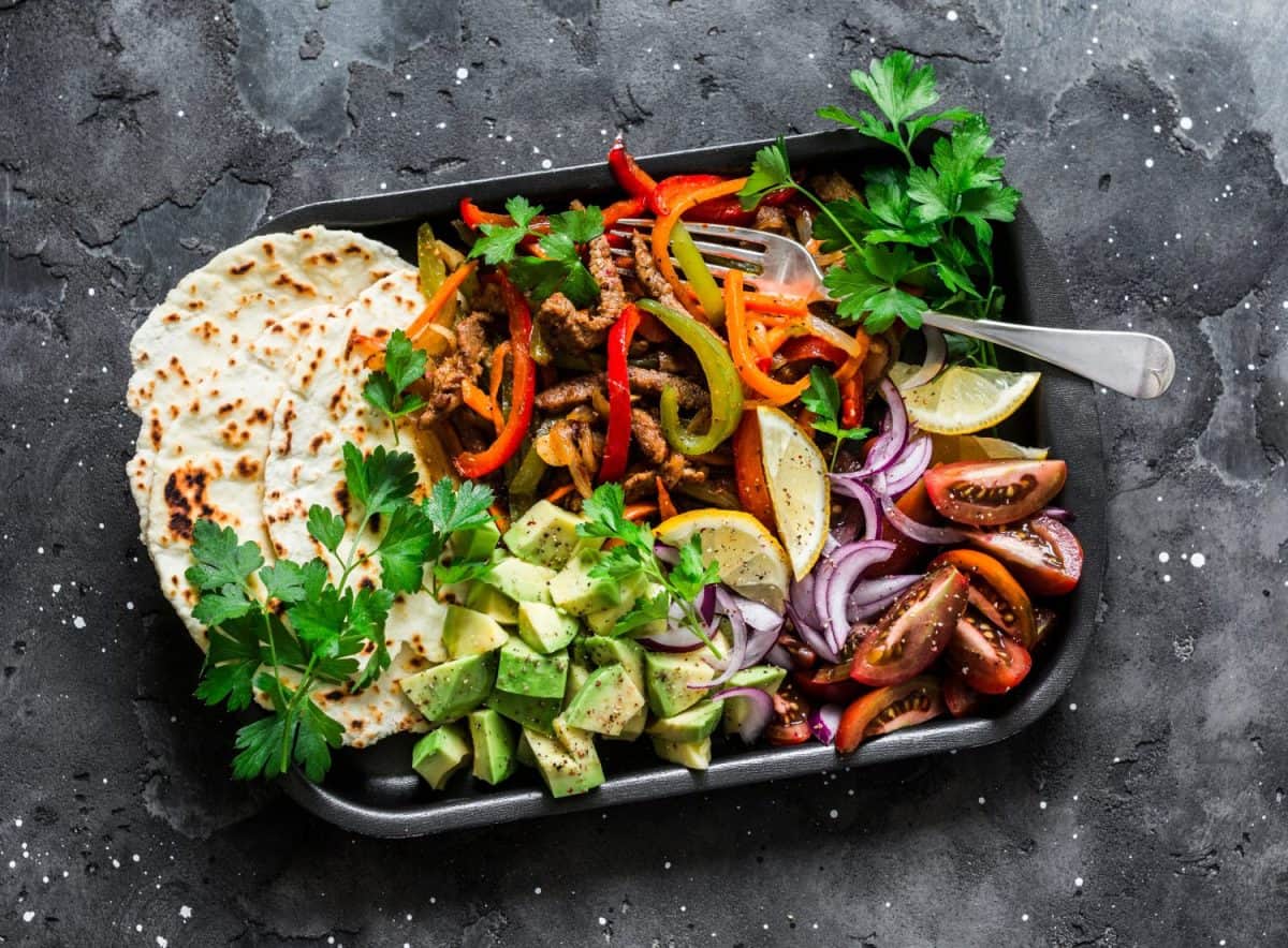 Spicy beef, vegetables, avocado, corn tortillas fajitas on a sheet pan on a dark background, top view. Delicious snack, tapas in mexican style. Sheet pan fajitas, ingredients, instructions and more