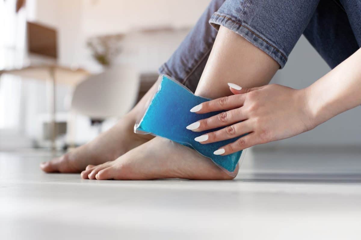 Close up woman feet and legs, and hands holding ice gel pack as cold compress on ankle due to stretching or injury, sitting on floor in apartment indoors. The best remedies for chigger bites.