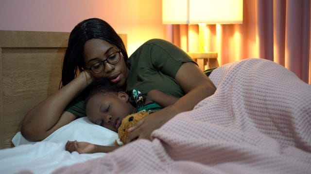 african american mother singing lullaby for little daughter before going to Sleep on bed at home. black mom story telling to girl in bedroom. sleeping child in parent embrace . kid hug Teddy bear. The best treatment for croup.