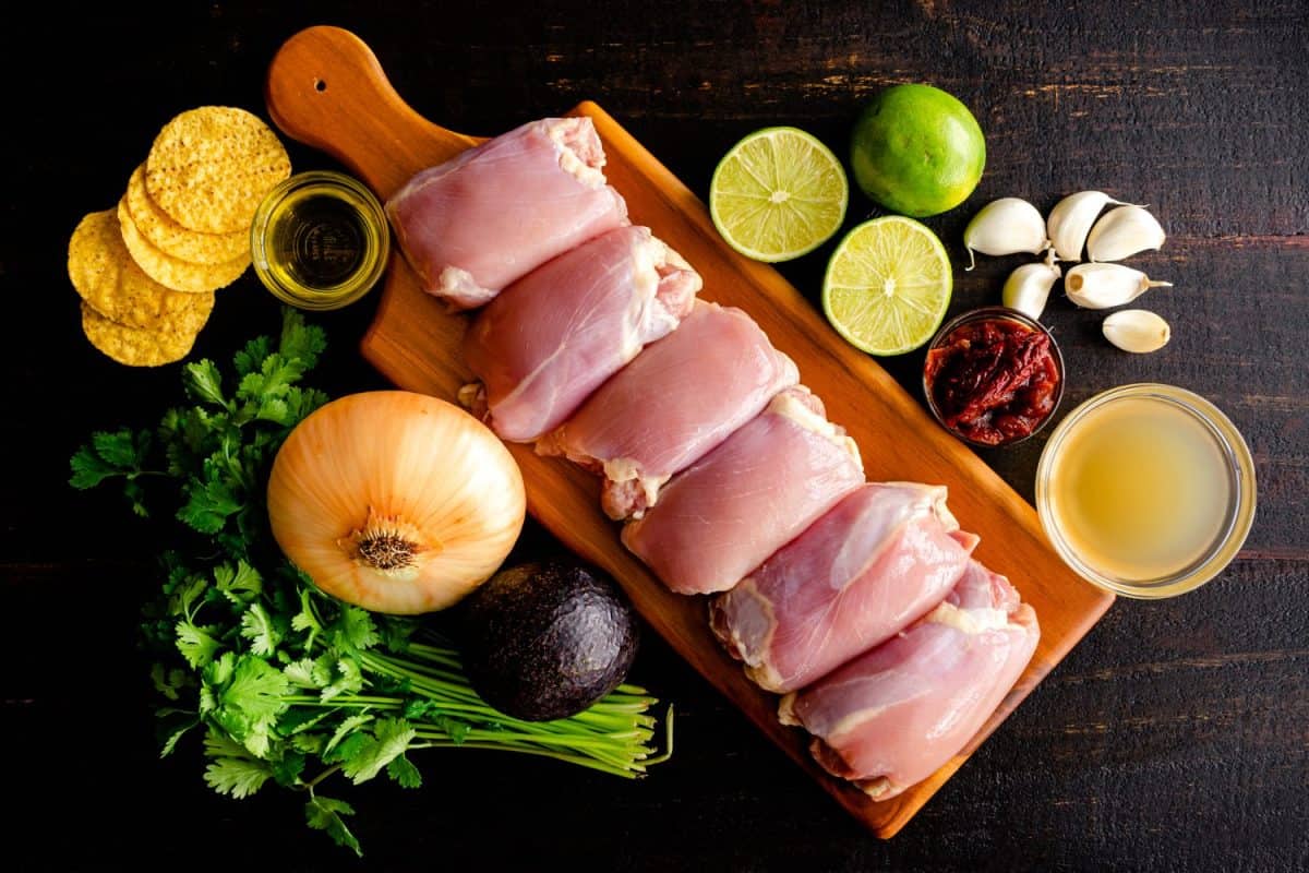 Mexican Chicken Lime Soup Ingredients on a Dark Wood Background: Raw chicken thighs, limes, chipotle peppers in adobo sauce, and other ingredients on a wood table. How to make sheet pan chicken thighs.