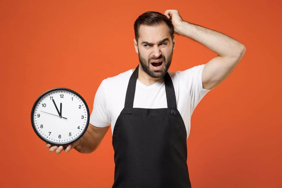 Young troubled disappointed man barista bartender barman employee in black apron white t-shirt work in coffee shop hold clock scratch head isolated on orange background Small business startup concept. Sheet Pan Meals: What They Are, How to Make Them