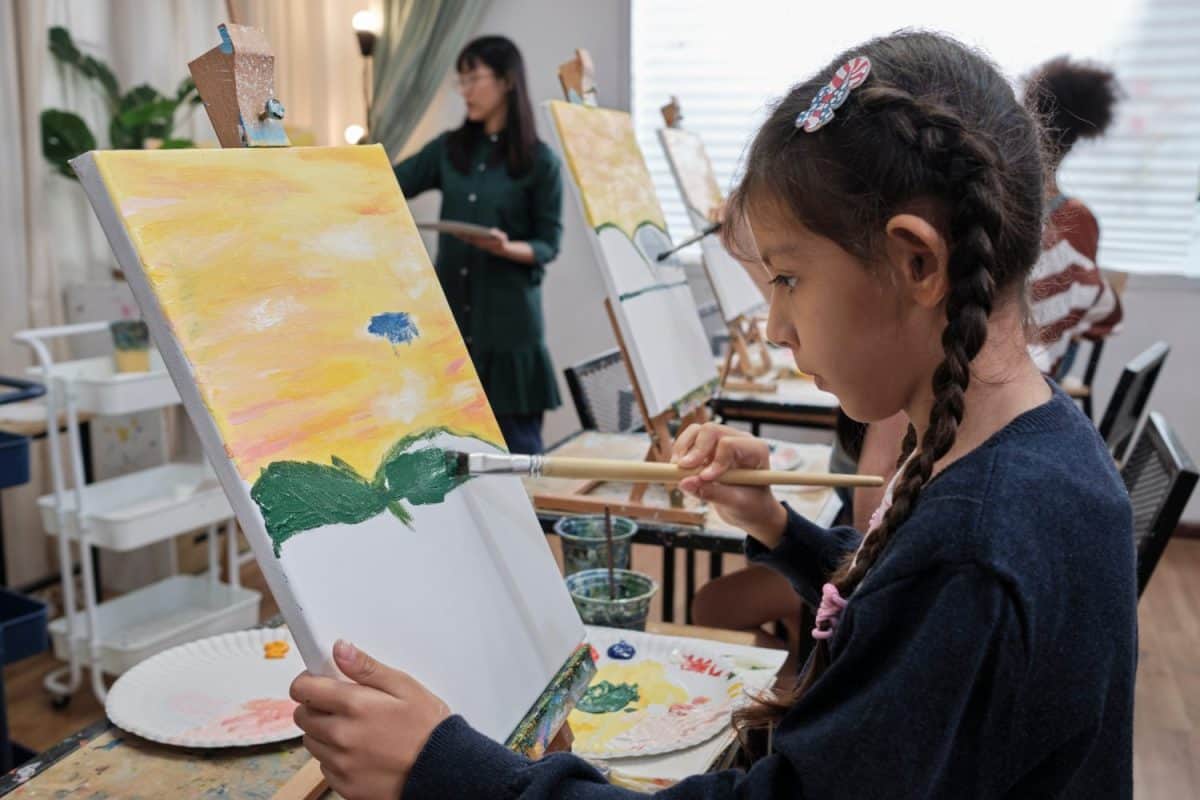 A little girl concentrates on acrylic color picture painting on canvas with student children in an art classroom, creative learning with talents and skills in the elementary school studio education. The best extra-curricular activities for creative kids.