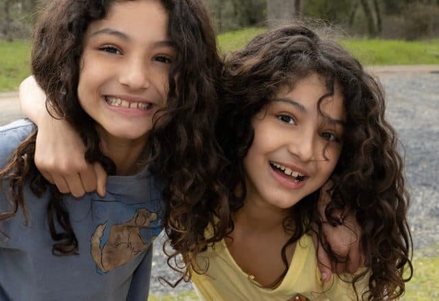 Pretty nine year old twin girls with curly hair rippling down their shoulders, laughing and hugging each other.