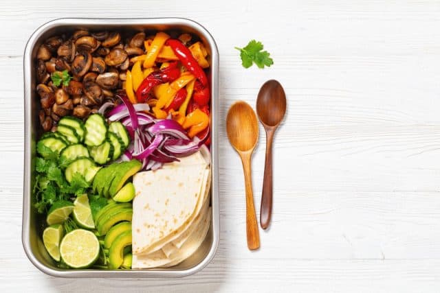 vegan fajitas with mushrooms, bell pepper, red onion, cucumber, avocado and cilantro in sheet pan with gluten free tortillas on white wooden table with spoons, flat lay, free space