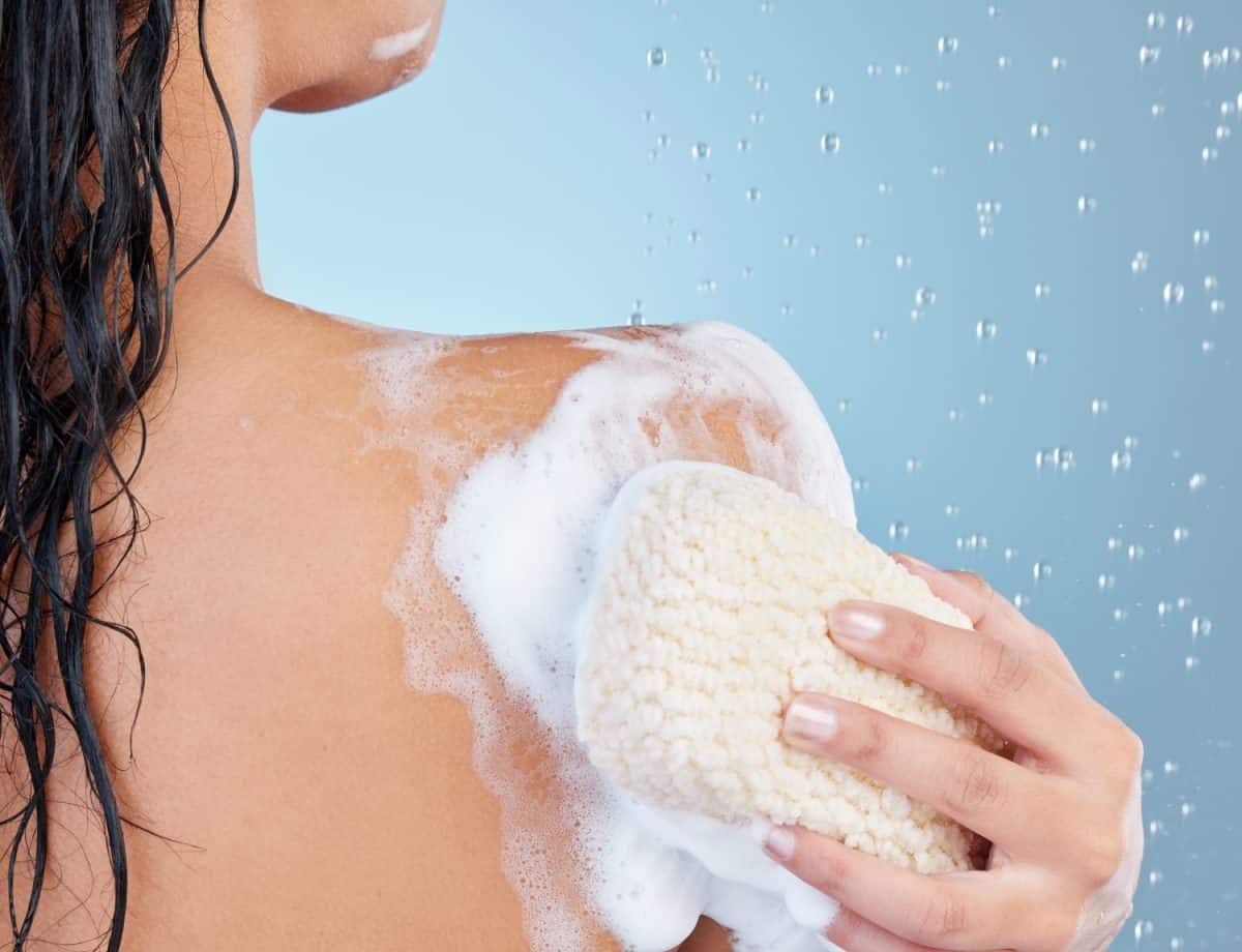 Hand, sponge and shower with soap in studio for hygiene, bacteria and dirt on back by blue background, Woman, person and model with loofah, foam or water drops with cleaning healthy body for skincare. The best remedies for chigger bites.