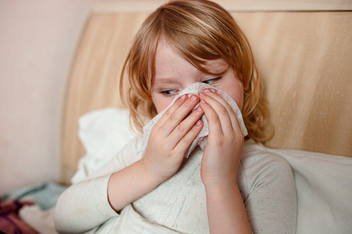 A young girl with long blonde hair lies in bed, blowing her nose into a tissue. She appears to be sick and is experiencing the common cold or flu symptoms.