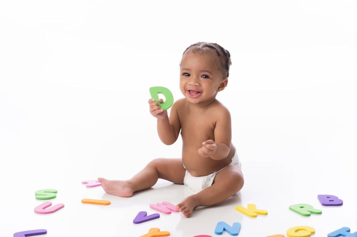 A smiling six month old baby girl wearing a white diaper. She is sitting on a white seamless background and playing with foam alphabet toys.