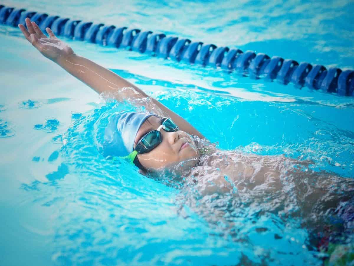 Kid swimming position backstroke. He put his hand up over the water. He wearing blue cap and goggle color black with a green strip.