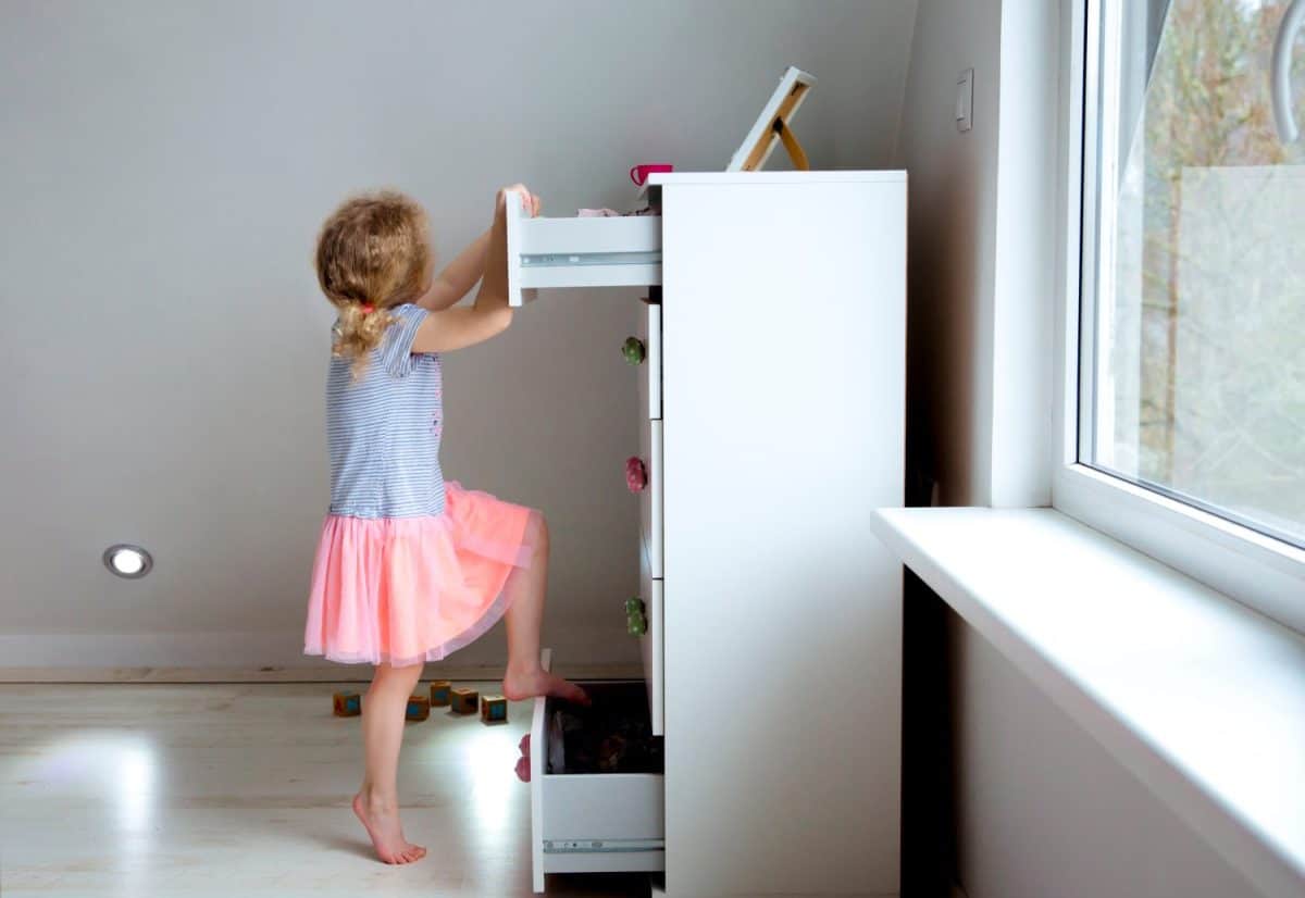 Young girl child climbing on modern high dresser furniture, danger of dresser dipping over concept. Children home hazards. Staged photo.