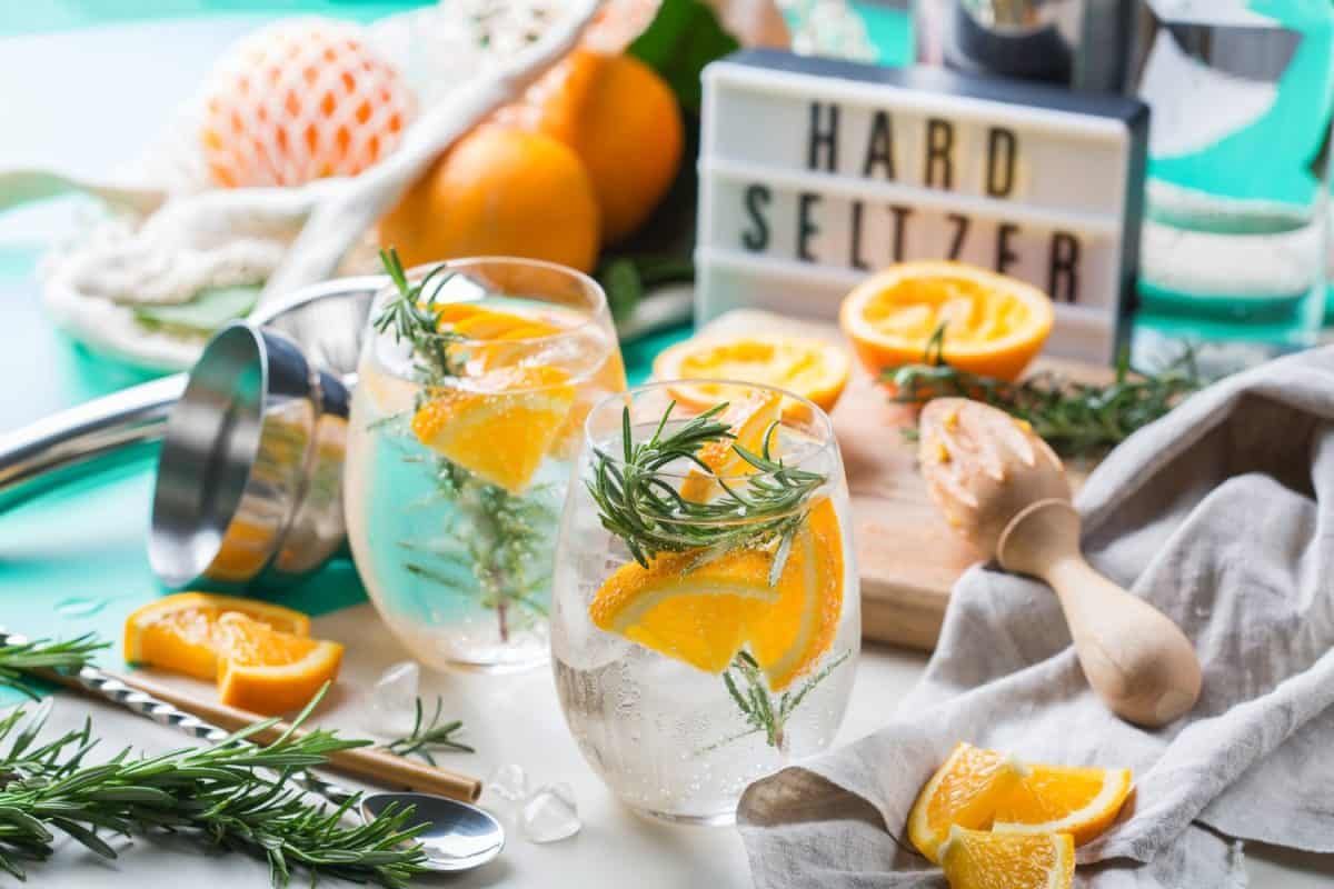 Hard seltzer cocktail with orange, rosemary and ice on a table. Summer refreshing beverage, drink with trendy zero waste accessories, bamboo straw and mesh bag.